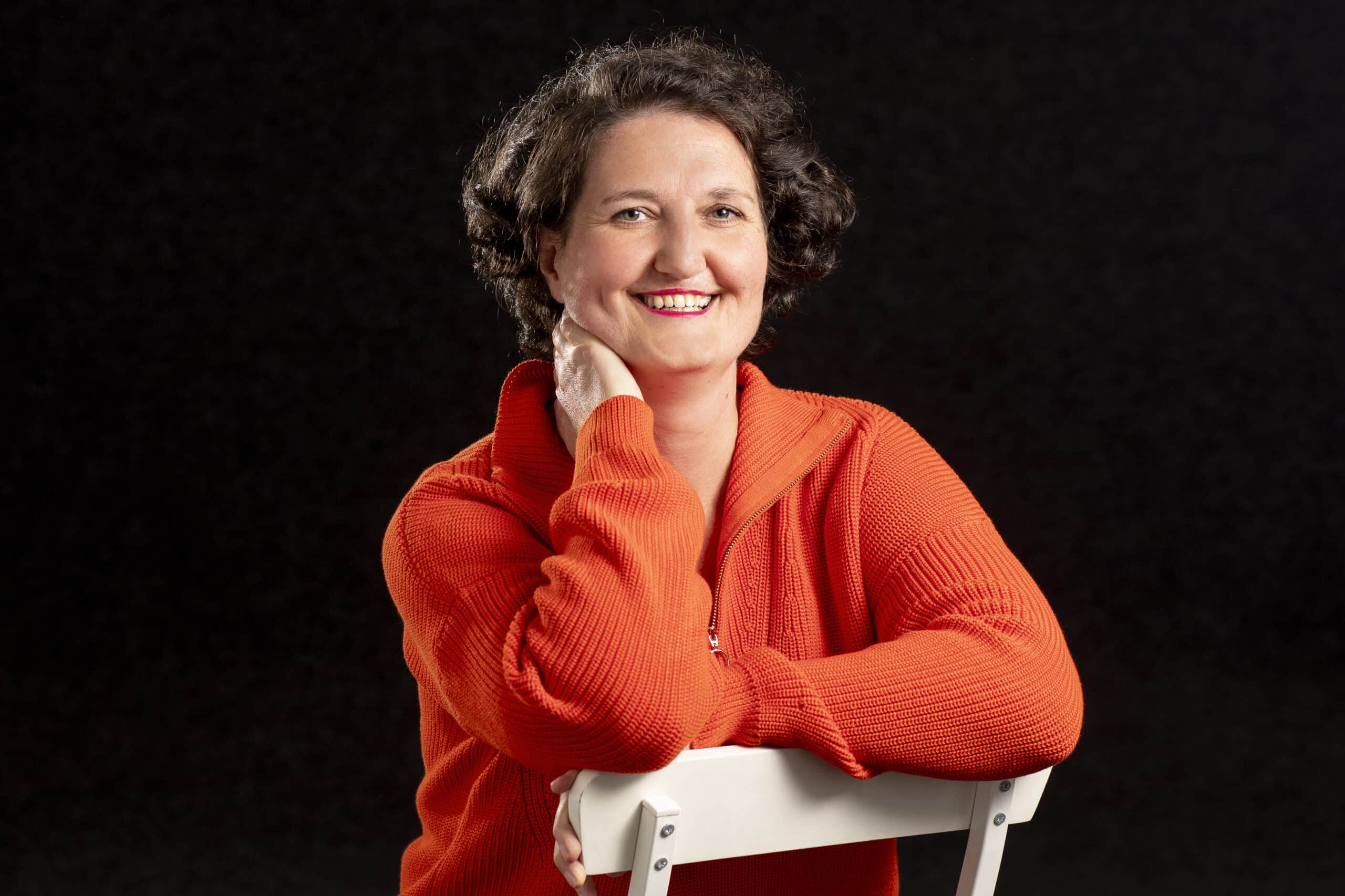Portrait_PersonalBrandingFotografin_AnnSophieDetje Personal Branding Fotografie Ann Sophie Detje Frau in einem orangen Pullover, schwarzer Hintergrund - lächeln im Gesicht.