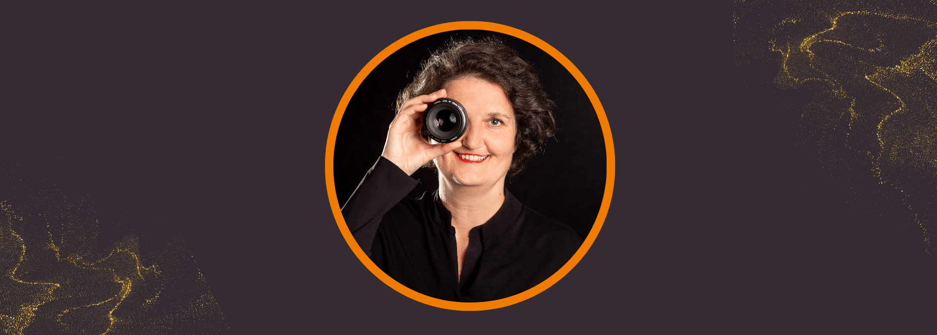 Blog-AnnSophieDetje-PersonalBranding-Fotografin_Markenbild Eine Frau mit lockigem, dunklem Haar und rotem Lippenstift hält ein Kameraobjektiv spielerisch vor ihr rechtes Auge und lächelt. Sie trägt Schwarz und ist kreisförmig eingerahmt von einem orangefarbenen Ring auf dunkelgrauem Hintergrund mit goldenen Akzenten. Bild von der mobilen Personal Branding Fotografin.