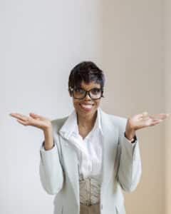 Fotoshooting für Gründer – Ann Sophie Detje – Personal Branding Fotografin: Eine Frau in hellem Business-Outfit mit Brille steht vor hellem Hintergrund. Sie lächelt und hebt beide Hände fragend und offen nach oben – eine Geste, die Leichtigkeit und Authentizität vermittelt.