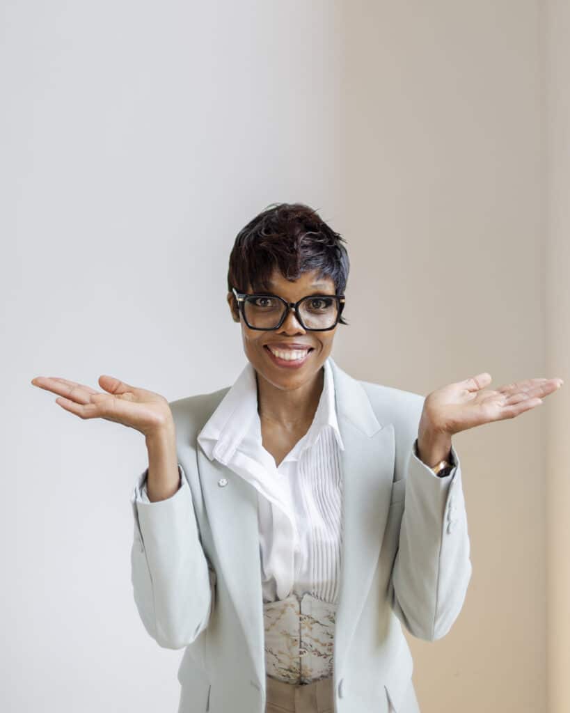 Fotoshooting für Gründer – Ann Sophie Detje – Personal Branding Fotografin: Eine Frau in hellem Business-Outfit mit Brille steht vor hellem Hintergrund. Sie lächelt und hebt beide Hände fragend und offen nach oben – eine Geste, die Leichtigkeit und Authentizität vermittelt.