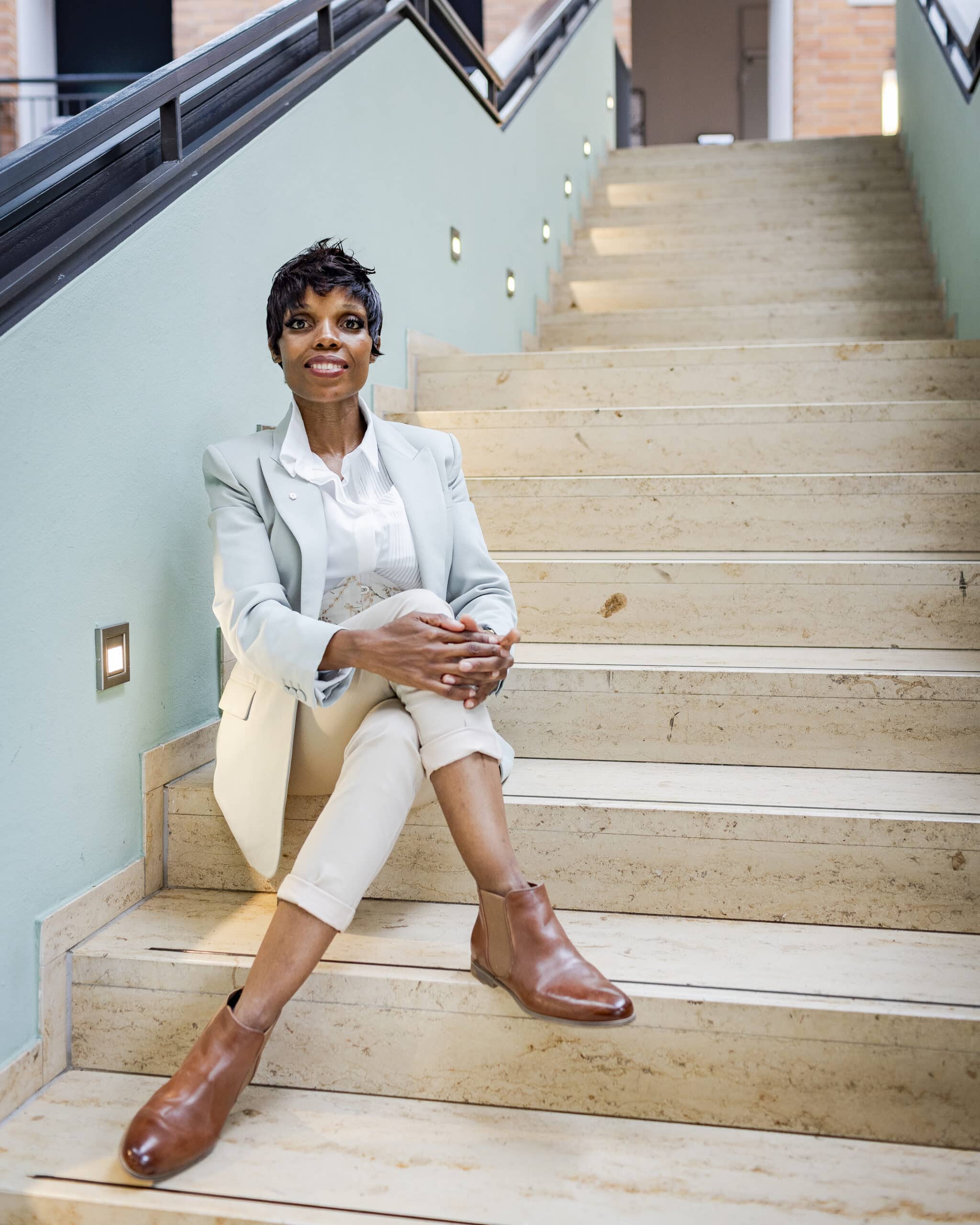 Fotoshooting für Gründer – Ann Sophie Detje – Personal Branding Fotografin: Eine Frau in hellem Blazer, weißer Bluse und beigen Hosen sitzt entspannt auf einer Steintreppe. Mit braunen Lederstiefeletten und selbstbewusstem Lächeln blickt sie in die Kamera.